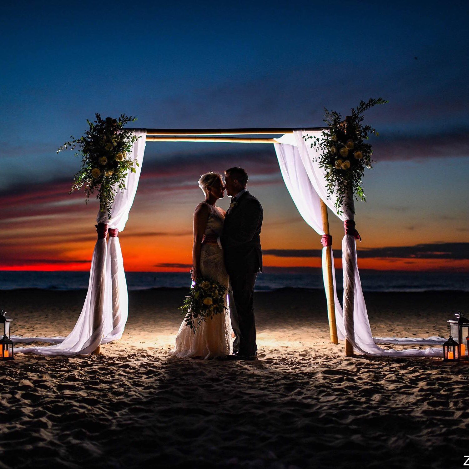 Couple standing on the beach under an arbor facing each other at sunset during the micro wedding